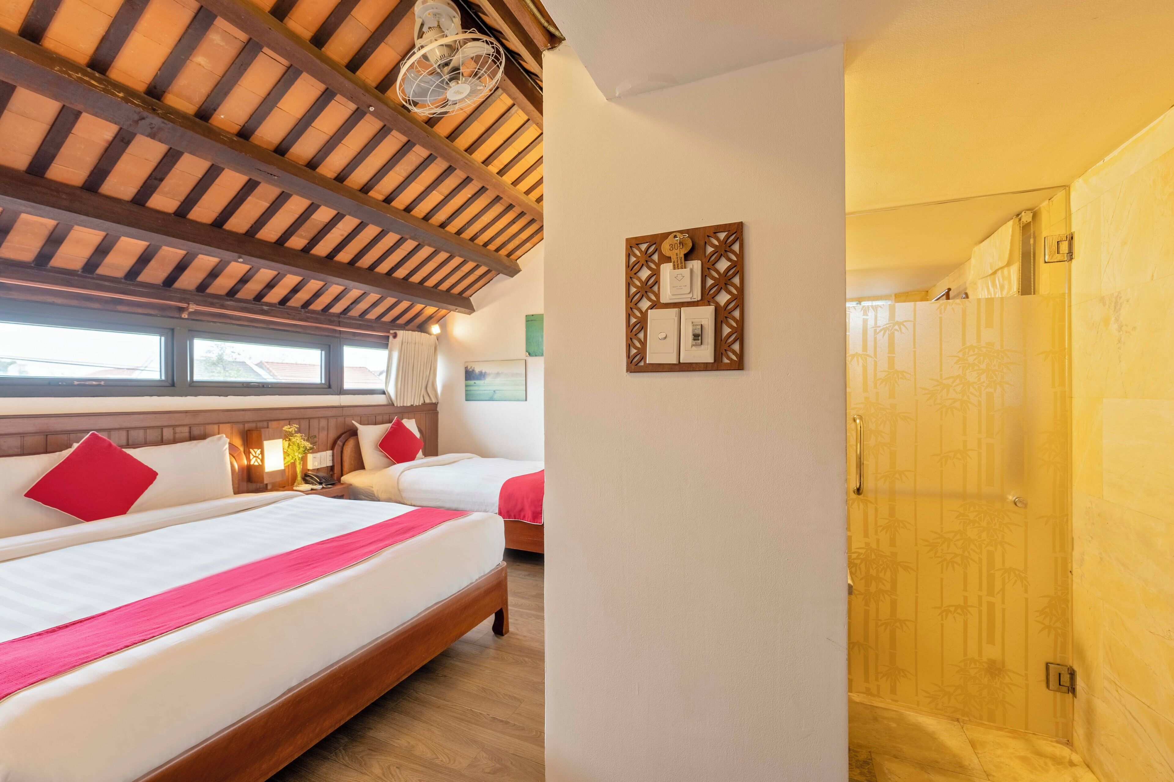 A hotel room with two beds, wooden ceiling beams, a wall-mounted light switch, and a shower area with bamboo wallpaper.