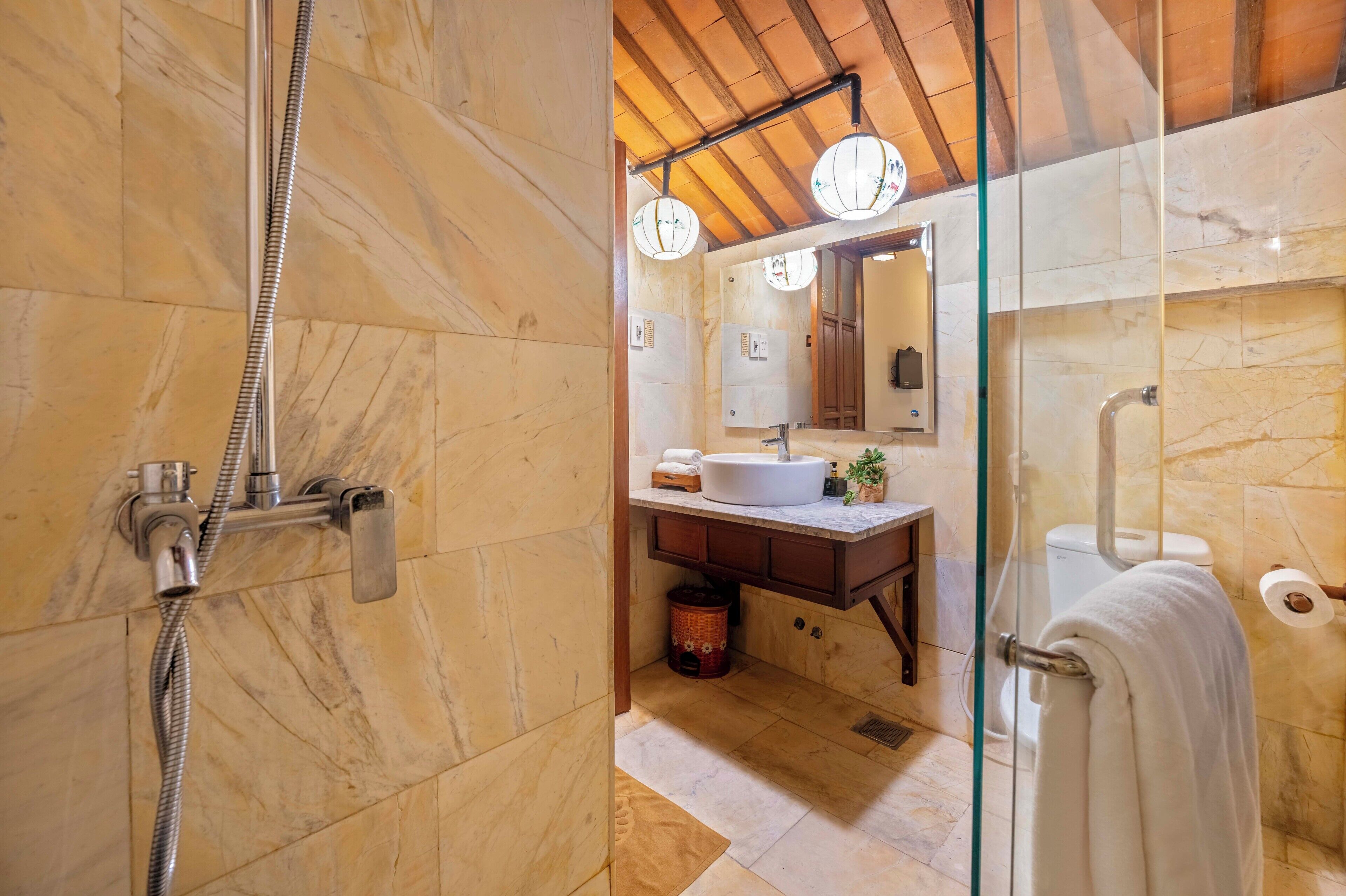 A bathroom with a glass-enclosed shower, marble tiles, a wooden ceiling, a sink, and a toilet with a towel hanging on the wall.