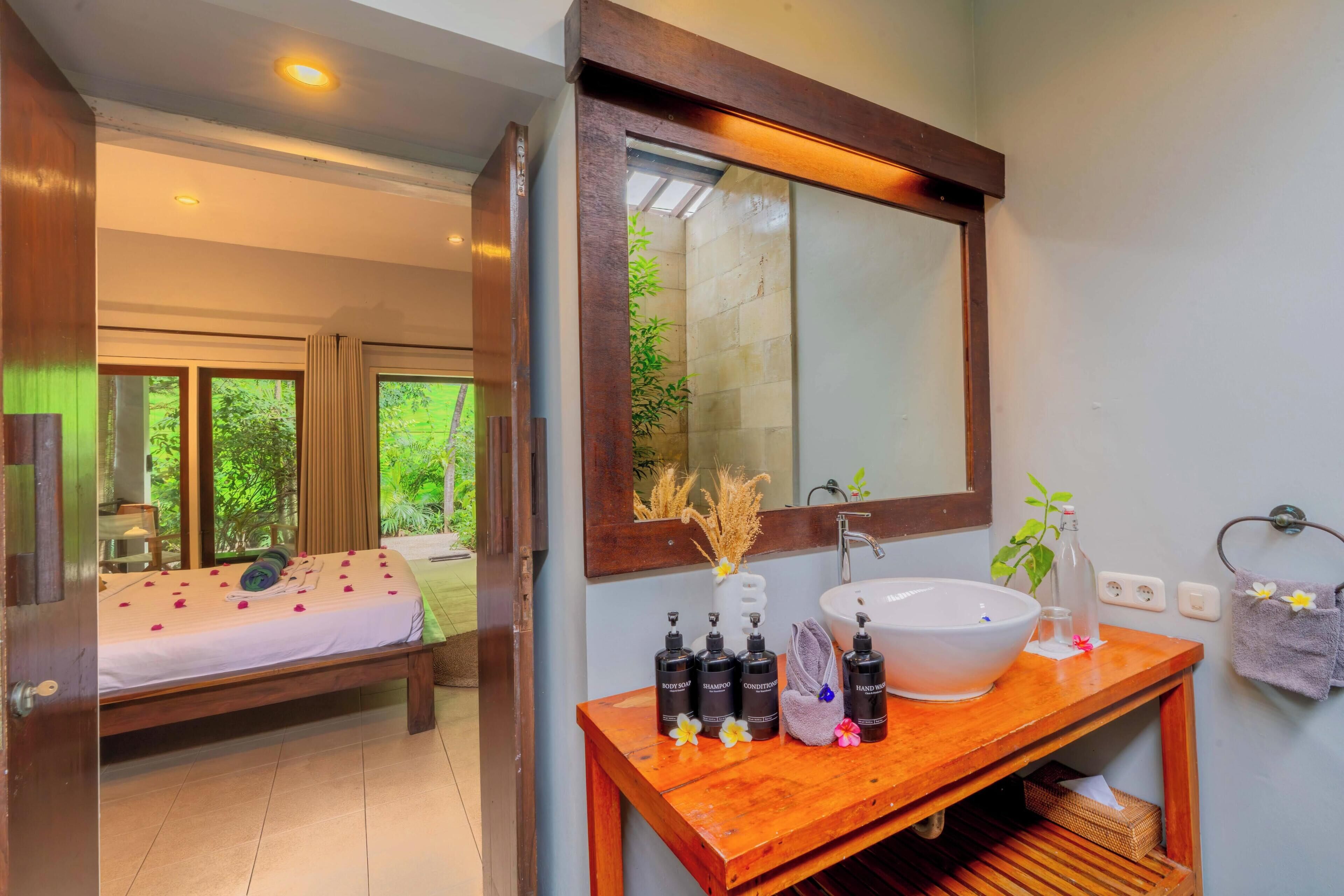 A bedroom with a bed, a wooden bedside table with toiletries, a large mirror, and a bathroom with a sink and towel rack.