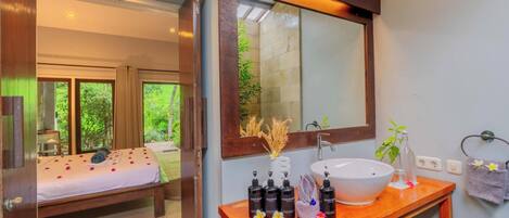 A bedroom with a bed, a wooden bedside table with toiletries, a large mirror, and a bathroom with a sink and towel rack.