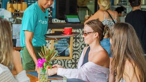 A bar setting with a person serving a drink, a table with a red cup, and a menu board in the background. - Bale Sampan Bungalows (Gili Trawangan)