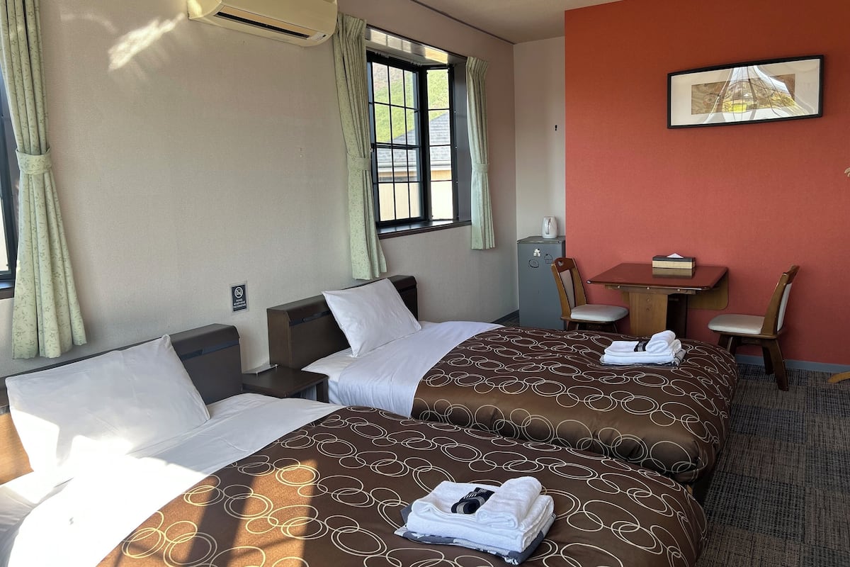 A hotel room with two beds, a desk, a chair, an air conditioning unit, and a window with curtains.