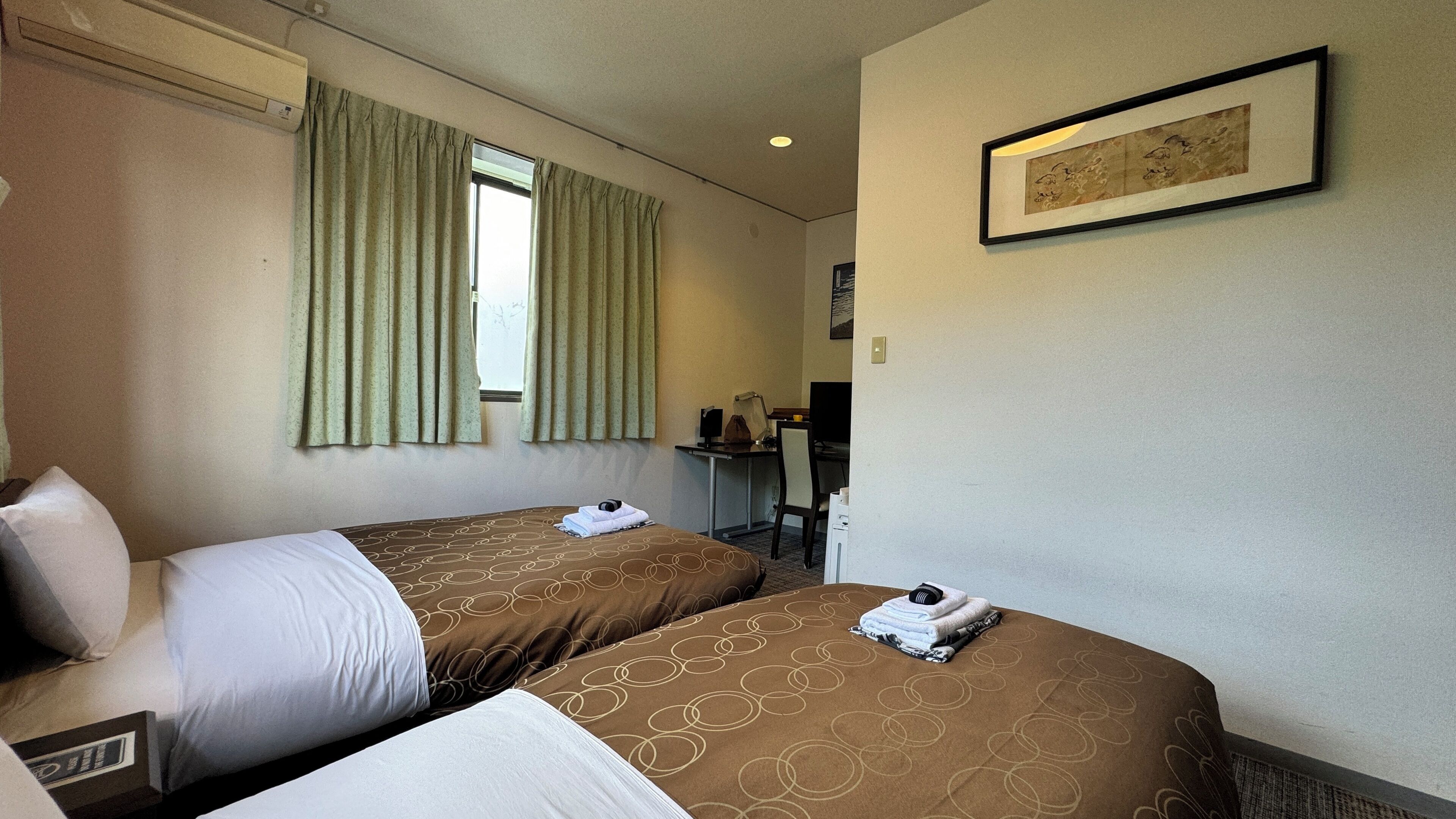 Habitación de hotel con dos camas, un escritorio, una silla, una ventana con cortinas y un cuadro en la pared.