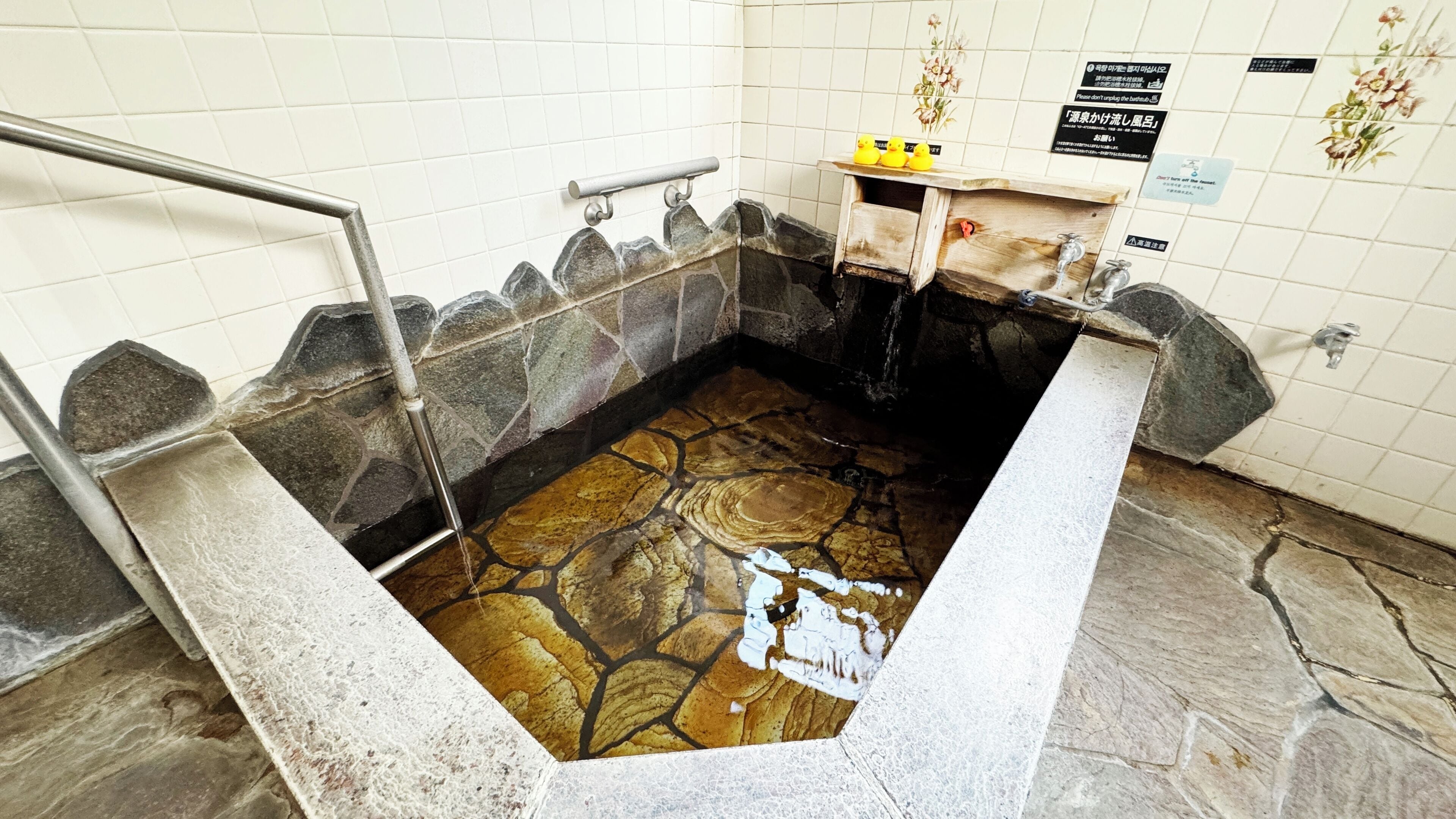 A stone hot spring bath with a metal railing, a wooden shelf with rubber ducks, and a tiled wall with signs.