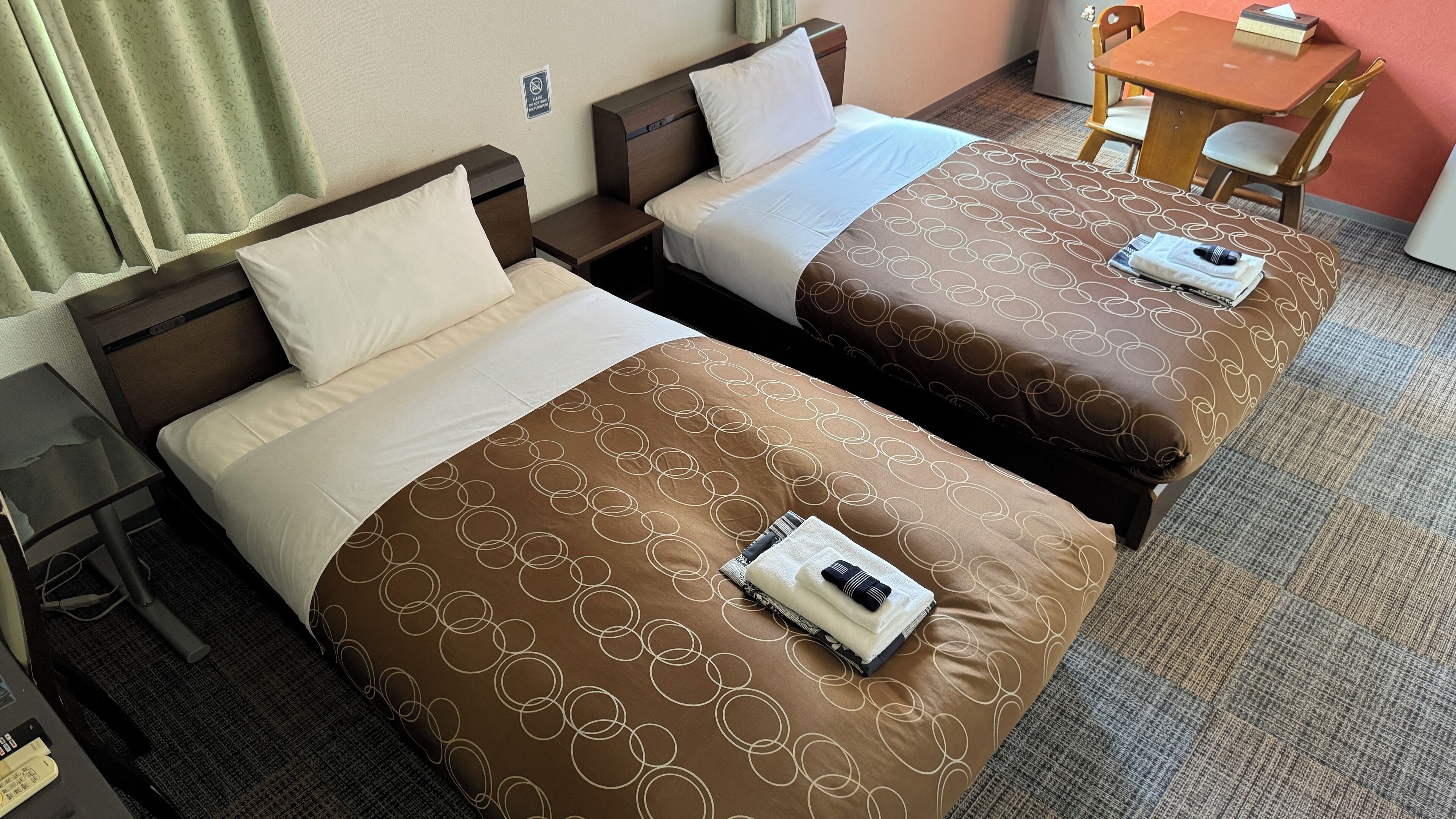 Dos camas individuales con ropa de cama marrón y blanca a juego, una mesita y una silla en una habitación de hotel.