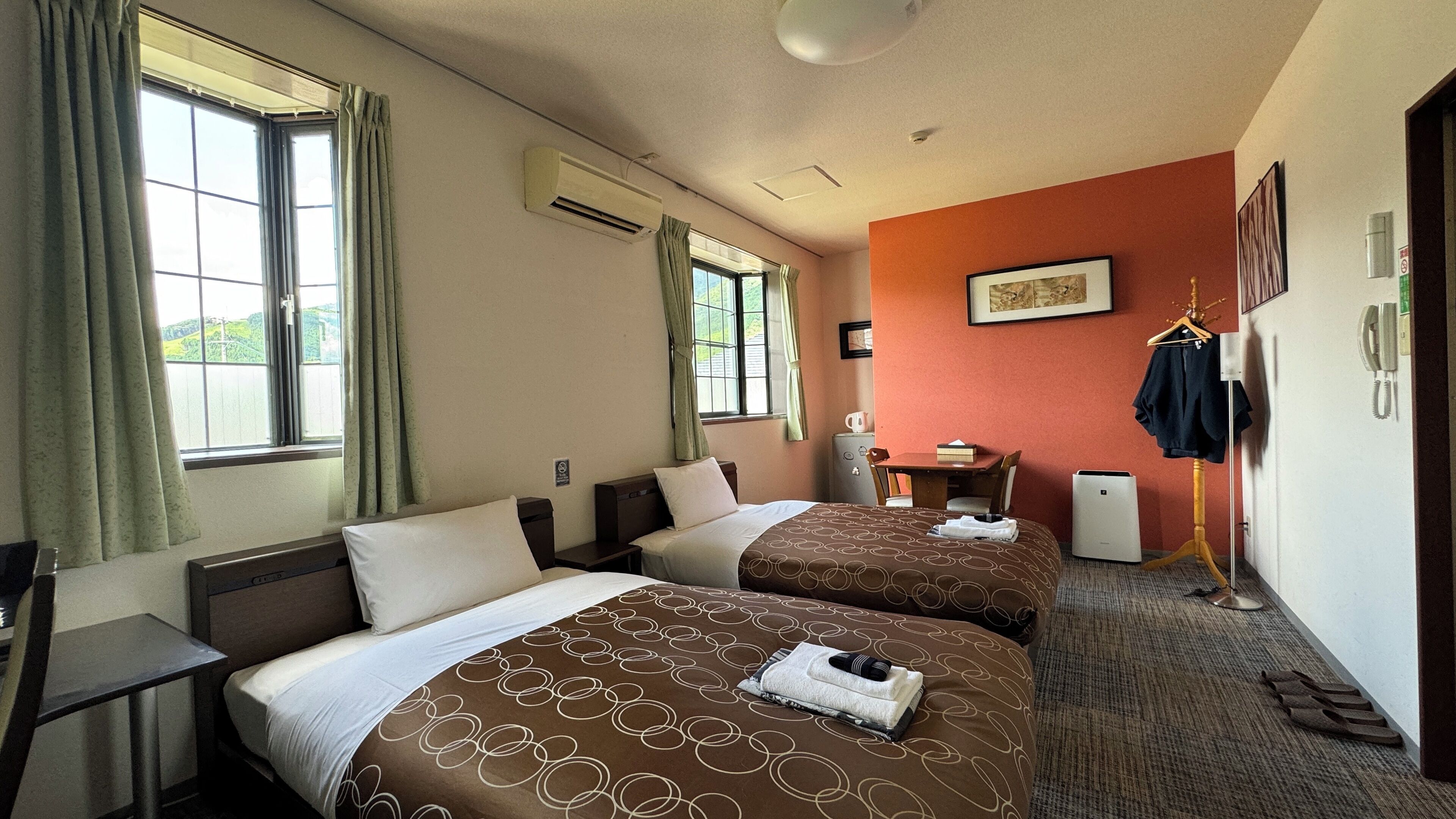 Ein Hotelzimmer mit zwei Betten, einem Schreibtisch, einem Stuhl, einem kleinen Tisch, einem Telefon und einem Kleiderhaken.