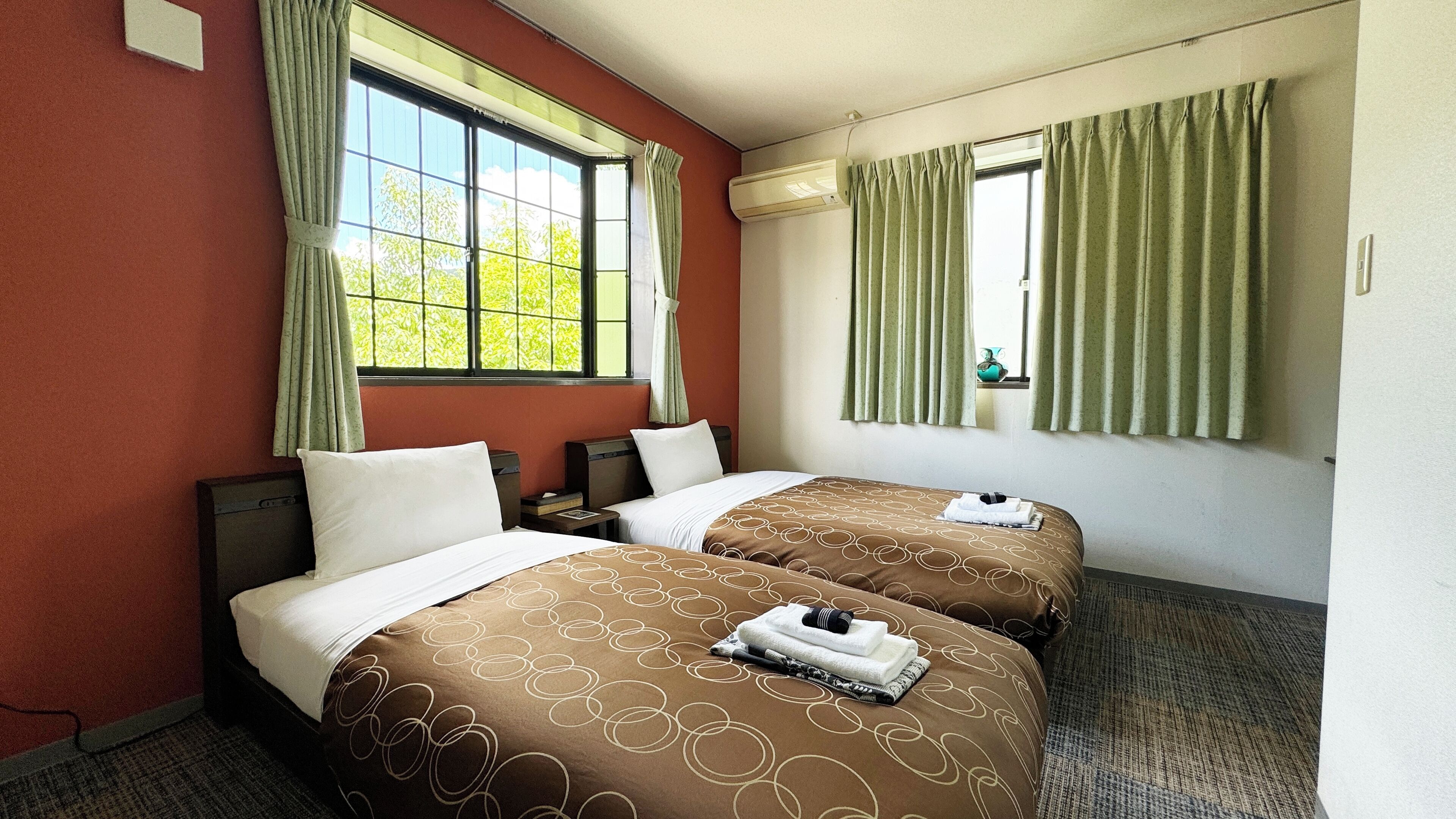 A hotel room with two beds, a window with curtains, an air conditioning unit, and a carpeted floor.