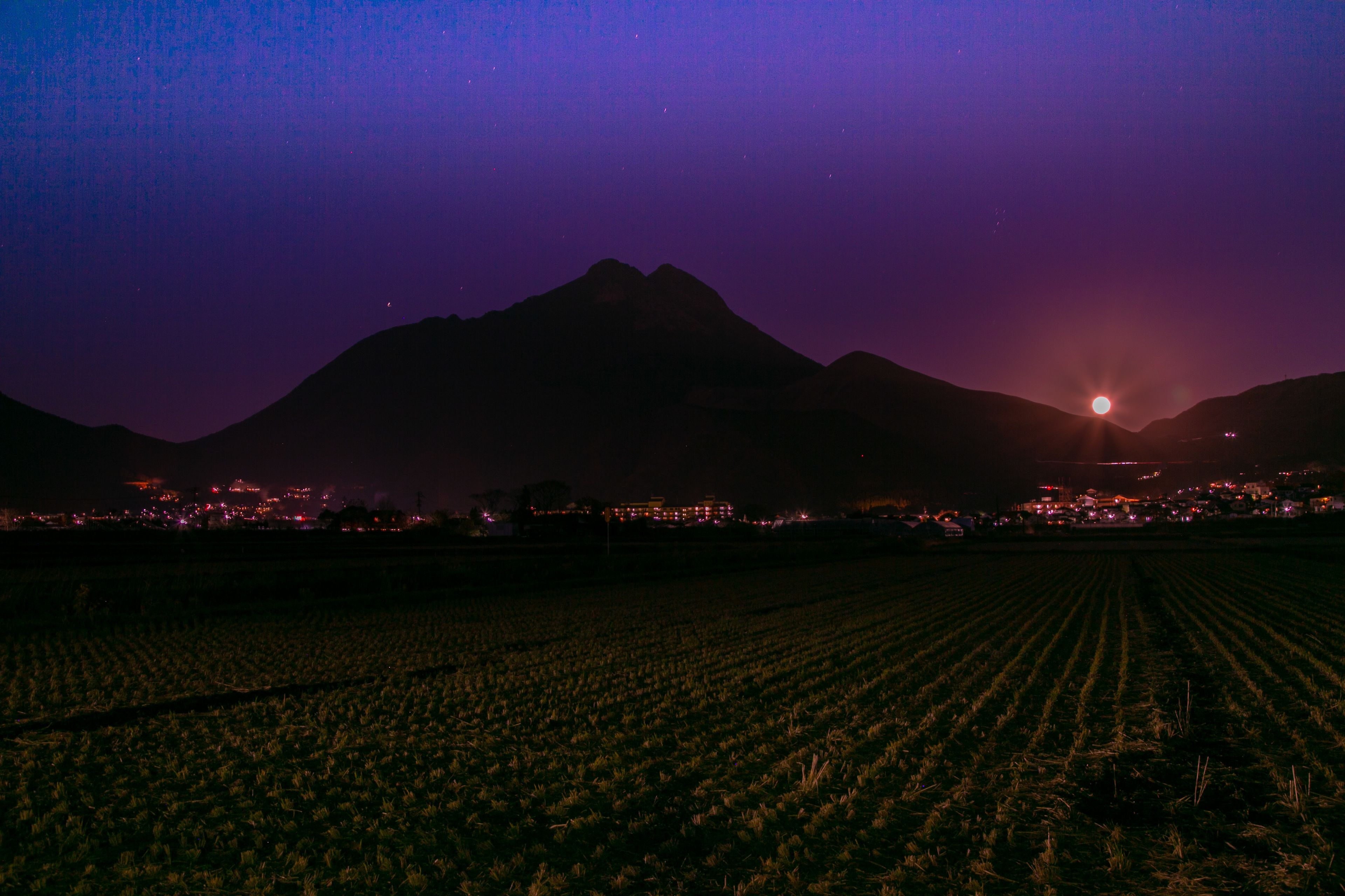 A nighttime landscape with a mountain silhouette, a field, and a distant cityscape with lights.