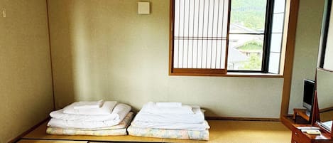 A traditional Japanese room with tatami flooring, two tatami mats, a futon bed, a television, and a window with a view of greenery.