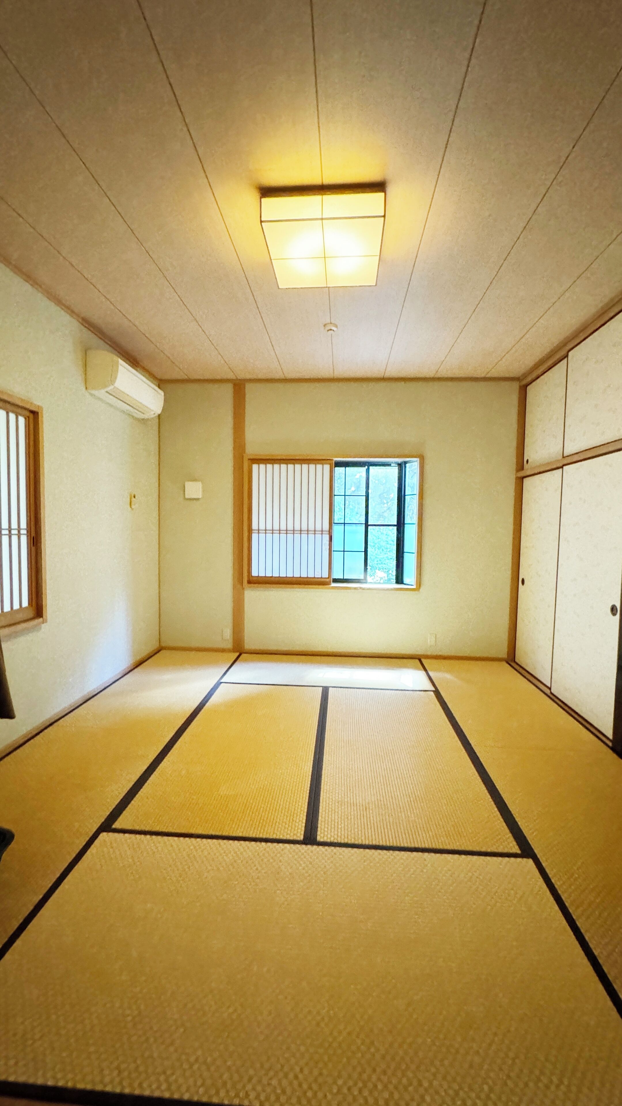Um quarto com tatames, uma porta deslizante, uma janela com persianas verticais e um aparelho de ar condicionado na parede.