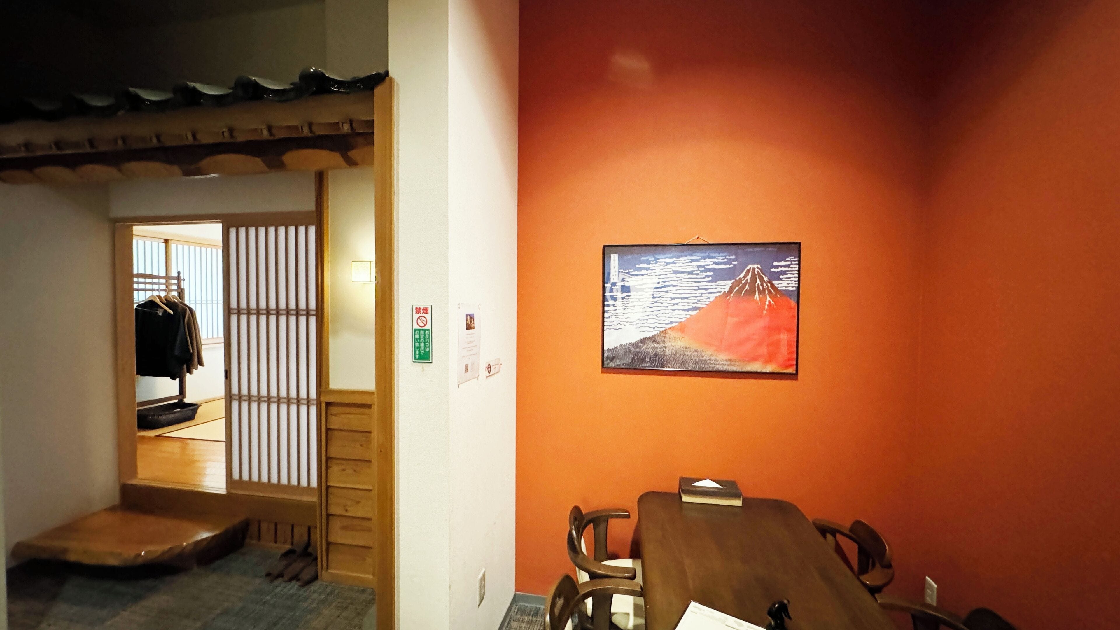 A room with a wooden table, chairs, a framed picture of a mountain, and a sliding door leading to a hallway with a coat rack and shoes.
