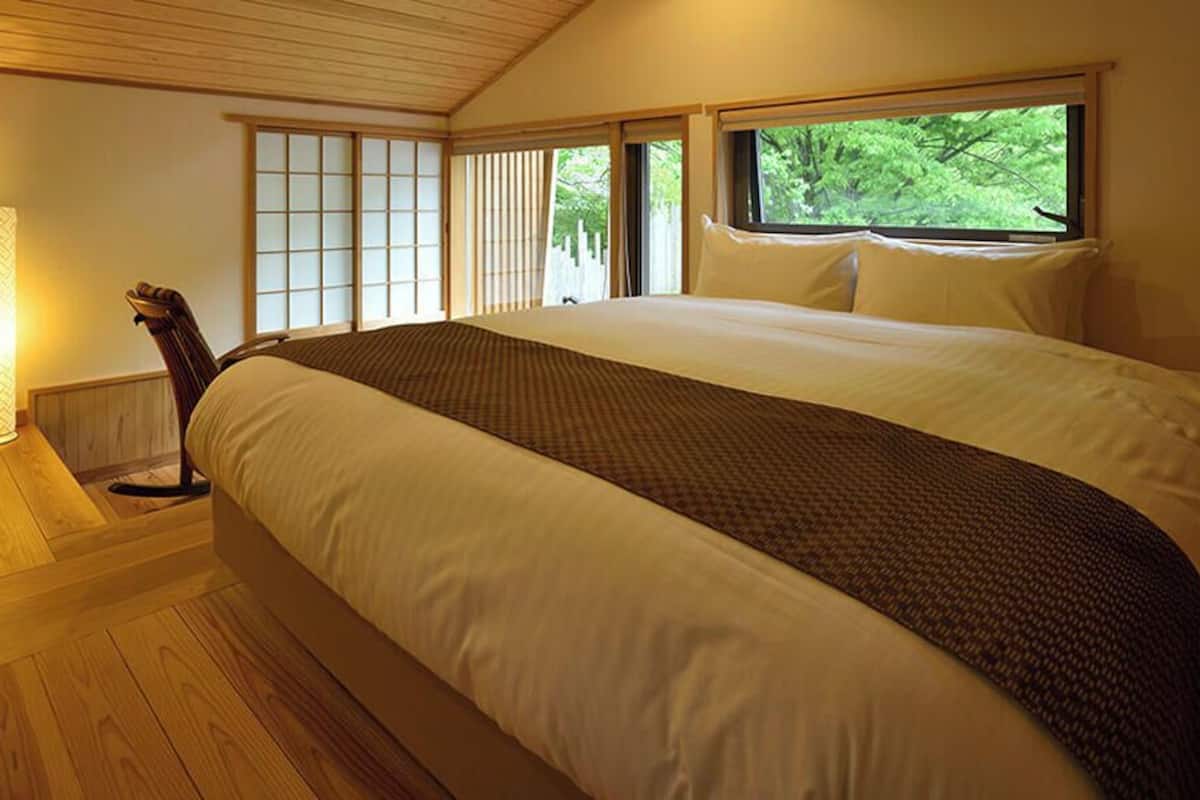 A bedroom with a large bed, a standing lamp, sliding doors, and a window with a view of greenery.