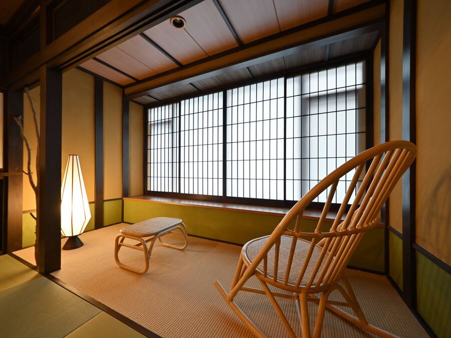 A traditional Japanese room with tatami flooring, a wooden chair, a small table, and a window with a grid pattern.