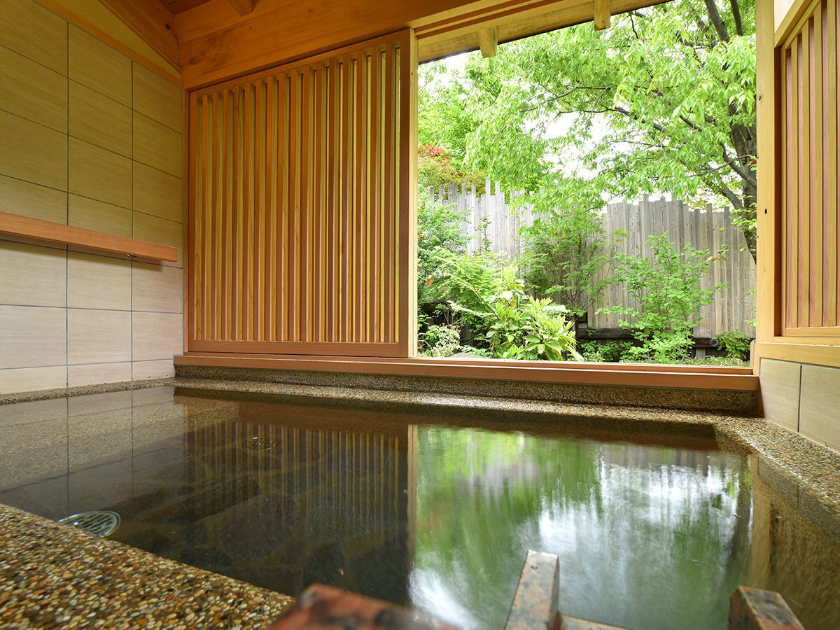 Un onsen japonés tradicional con interior de madera, un baño de piedra y vistas a una valla de madera y vegetación exterior.