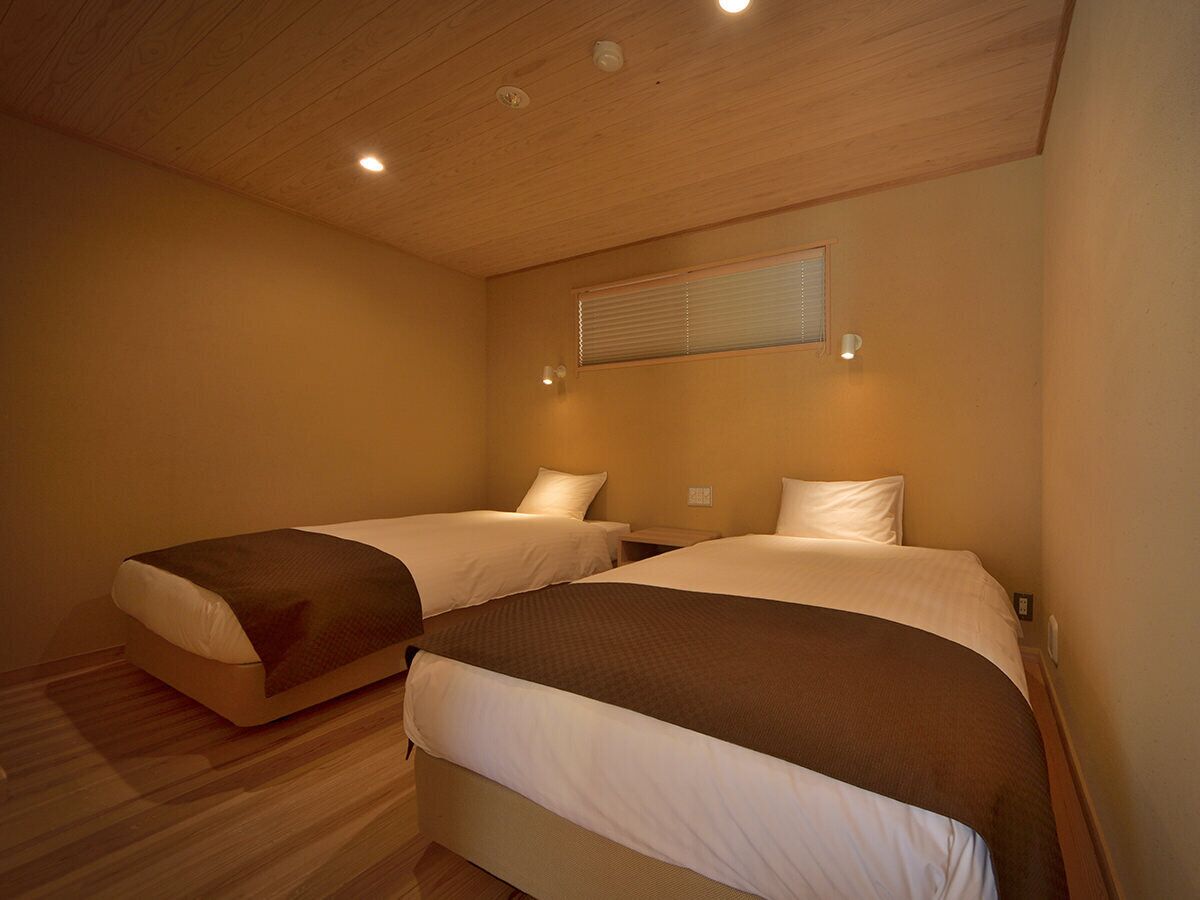 A room with two beds, wooden flooring, a ceiling light, and a window with blinds.