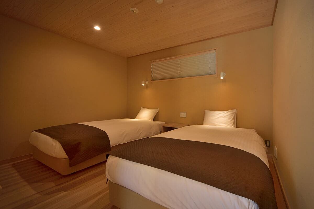 A room with two beds, wooden flooring, a ceiling light, and a window with blinds.