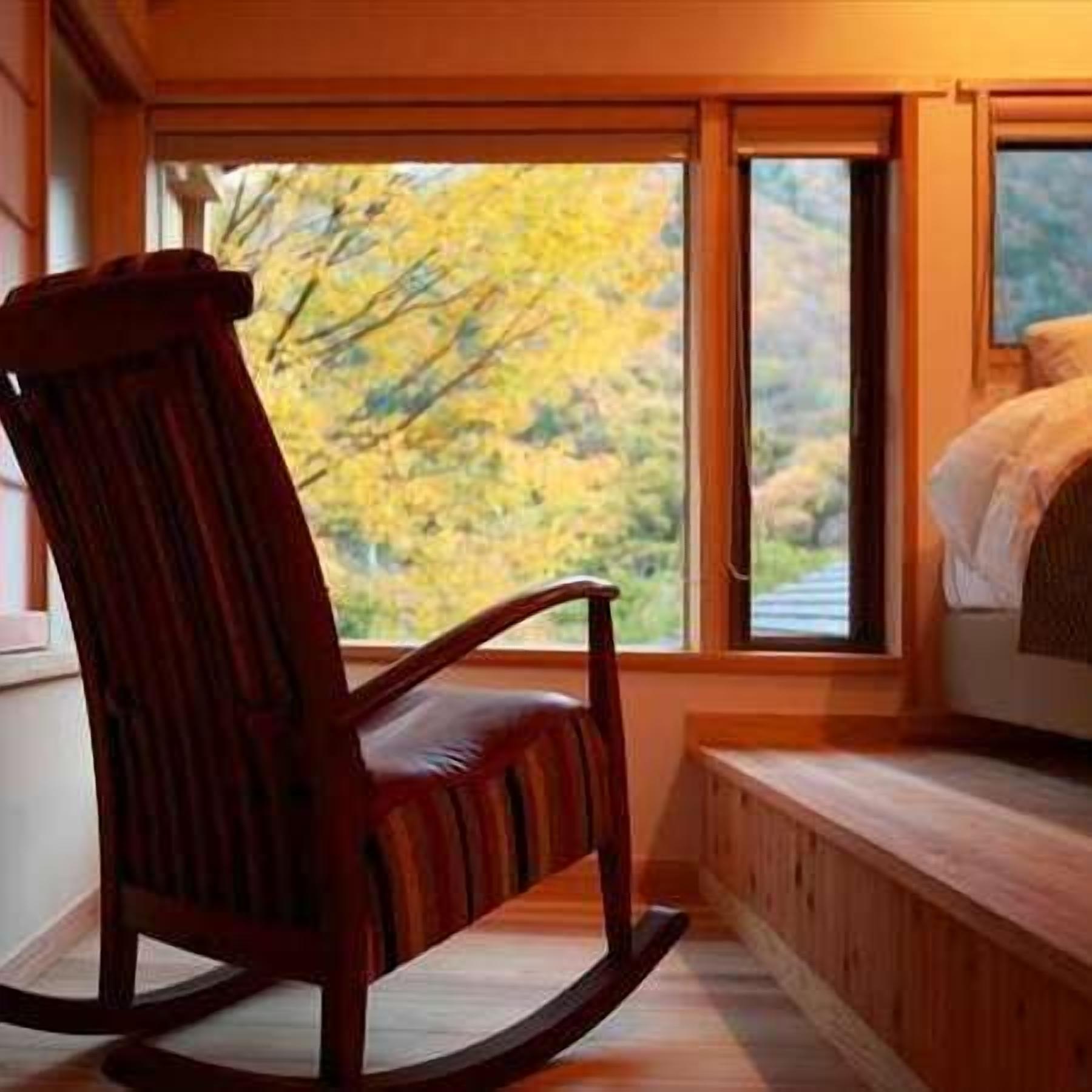 A rocking chair near a window with a view of autumn trees.