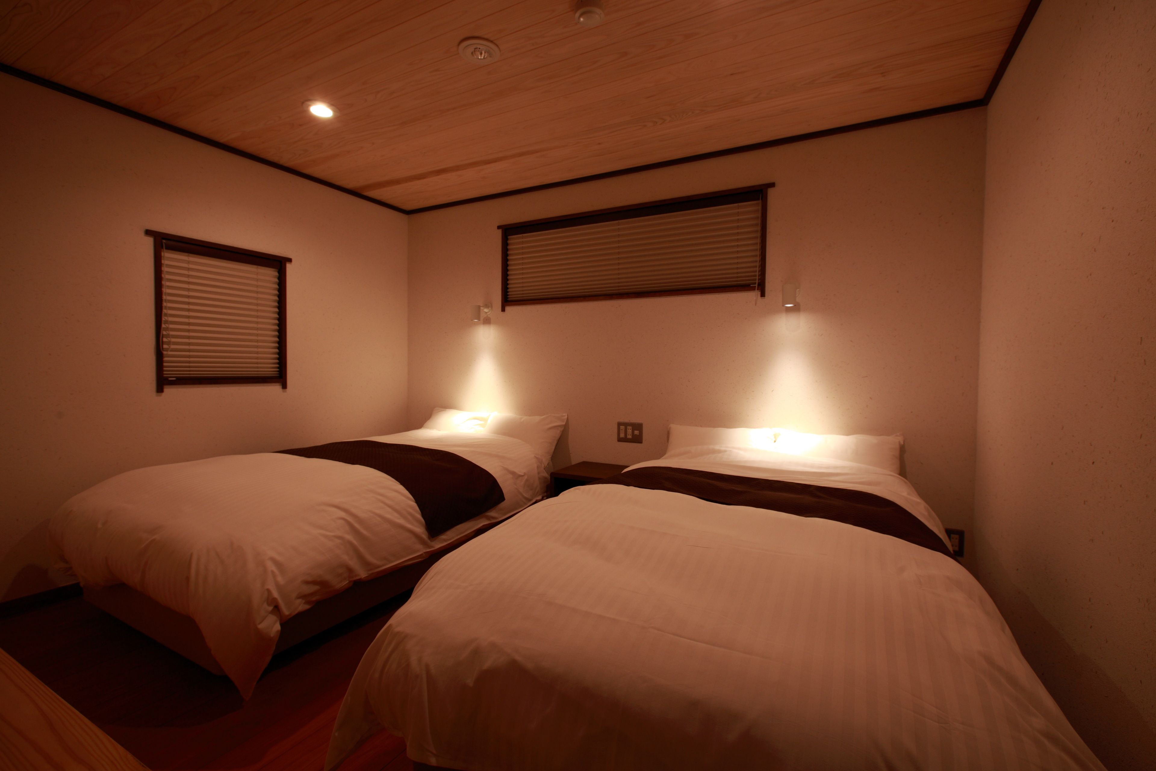 Quarto com duas camas, teto de madeira e duas luminárias fixadas na parede.
