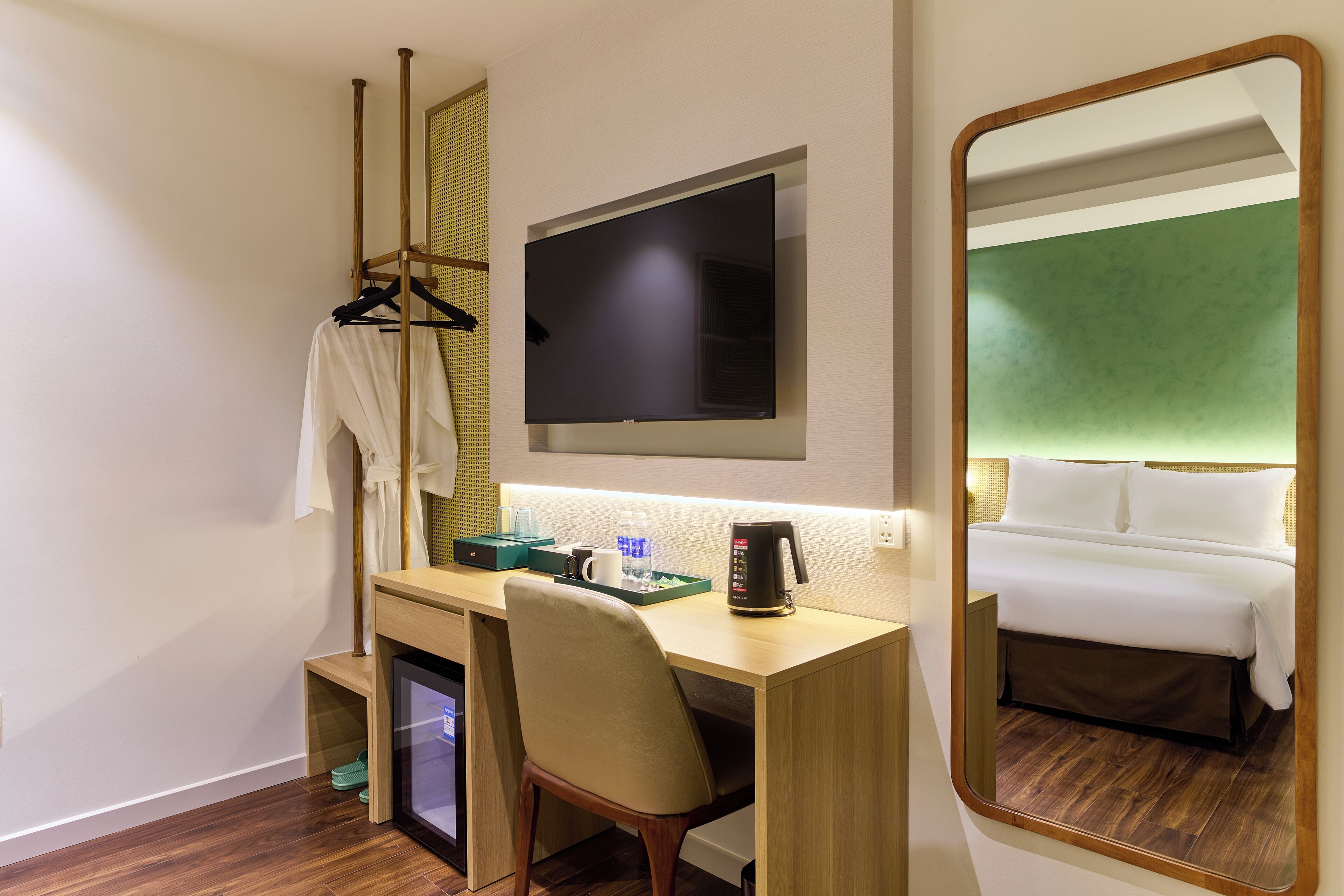 A hotel room with a wooden desk, a television mounted above it, a mirror reflecting the room, a bed with white bedding, and a small desk area with a coffee maker and a tray with glasses and water bottles.