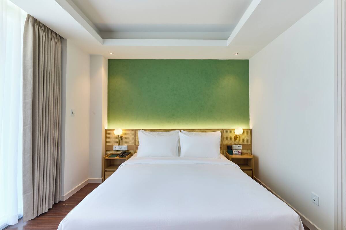 Une chambre d’hôtel avec un grand lit, deux tables de chevet, un mur d’accent vert et une fenêtre avec des rideaux.