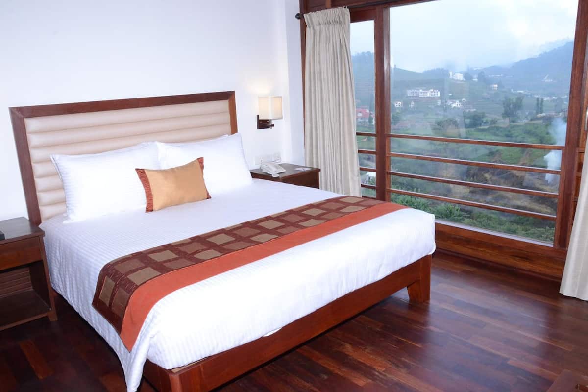 A hotel room with a large bed, wooden headboard, a nightstand, a bedside table with a lamp, and a view of a mountainous landscape through a large window.
