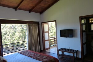 A bedroom with a large bed, a wooden bench, a TV mounted on the wall, and a balcony with a view of trees.