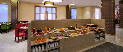 A buffet setup with various food items and beverages, a seating area with chairs, and a decorative wall with a floral pattern.
