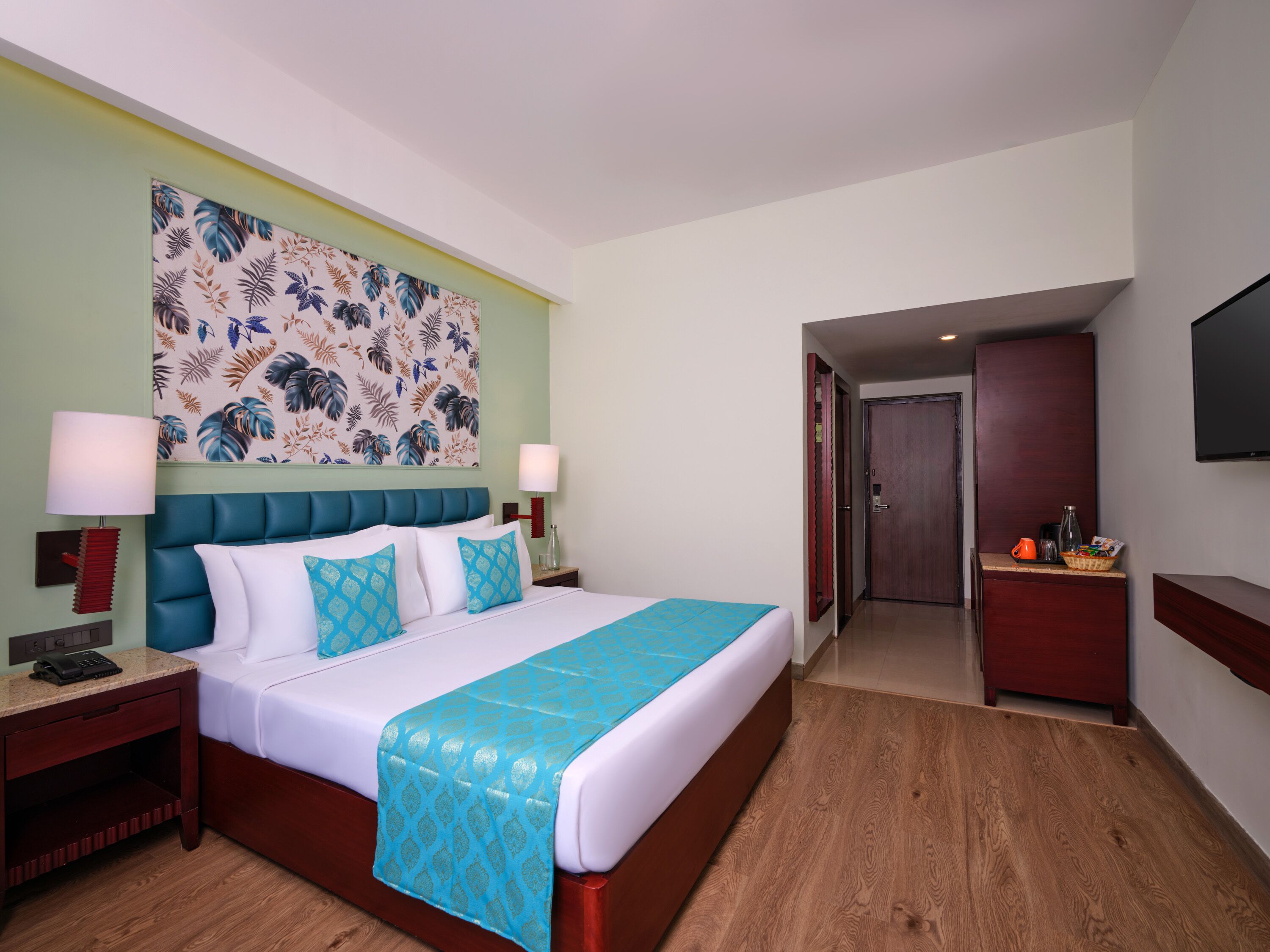 A hotel room with a bed, nightstands, a wall-mounted TV, and a door leading to another room.