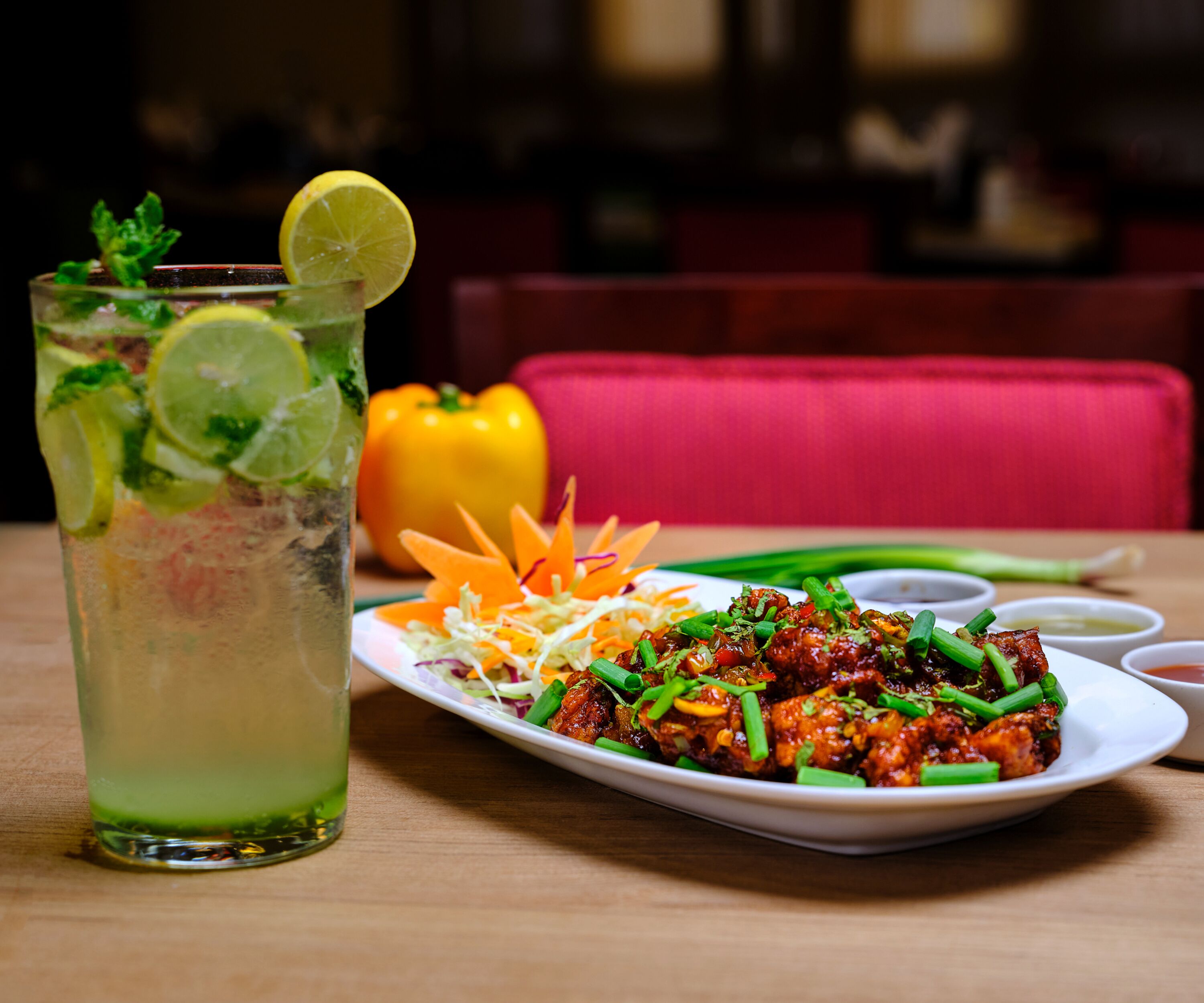A table with a glass of iced beverage garnished with lime and mint, a plate of stir-fried chicken with vegetables, and a yellow bell pepper in the background.