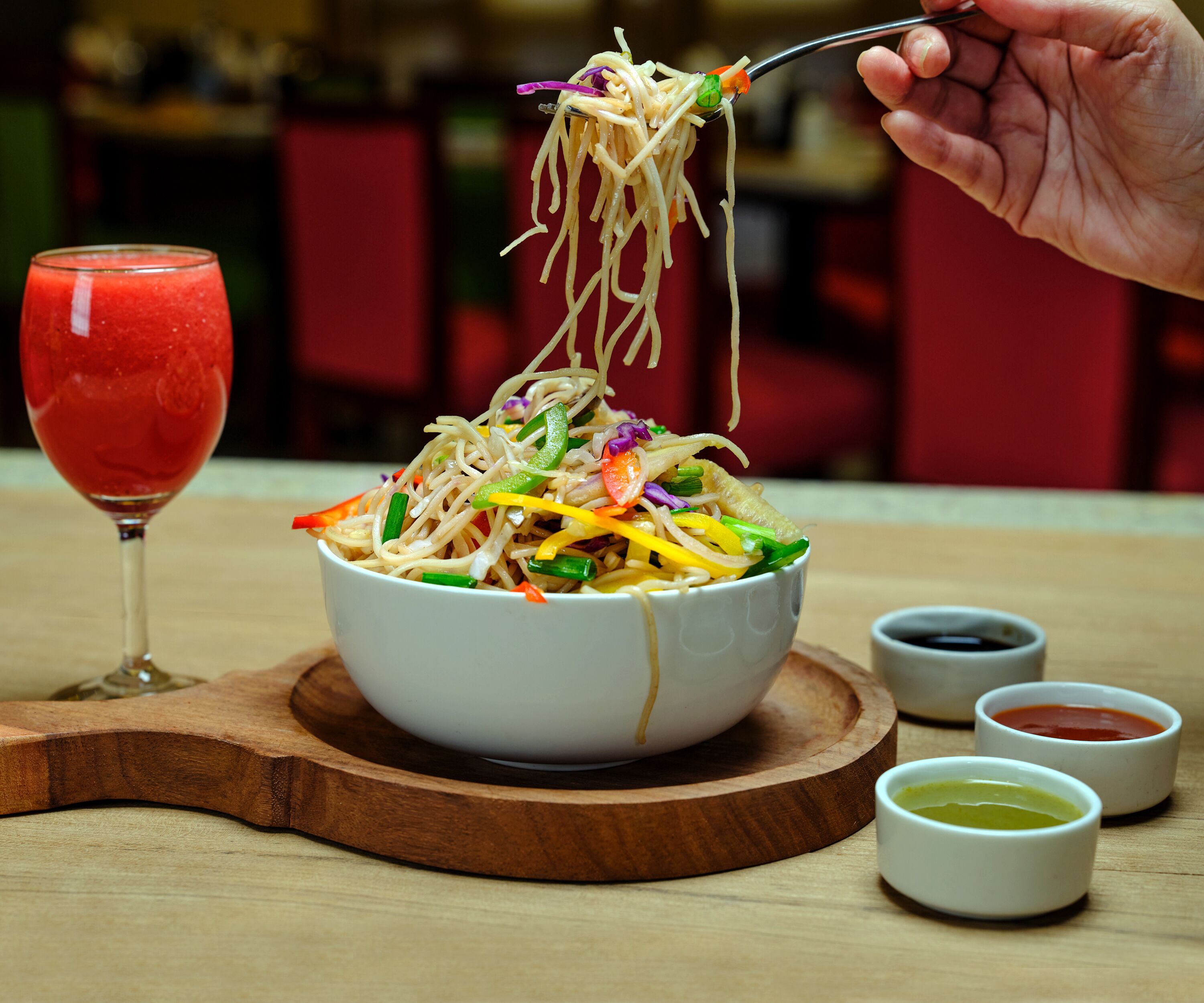 A bowl of noodles with vegetables and a glass of red drink on a wooden board.