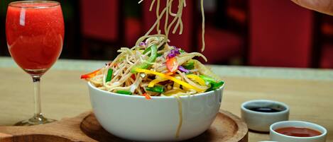 A bowl of noodles with vegetables and a glass of red drink on a wooden board.