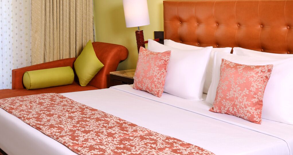 A hotel room with a large bed, a red upholstered chair, and a nightstand with a lamp.