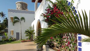 A building with a stone tower, arched doorways, and a garden with palm trees and flowering plants. - Squares Cafe and Apartments (Puerto Galera)