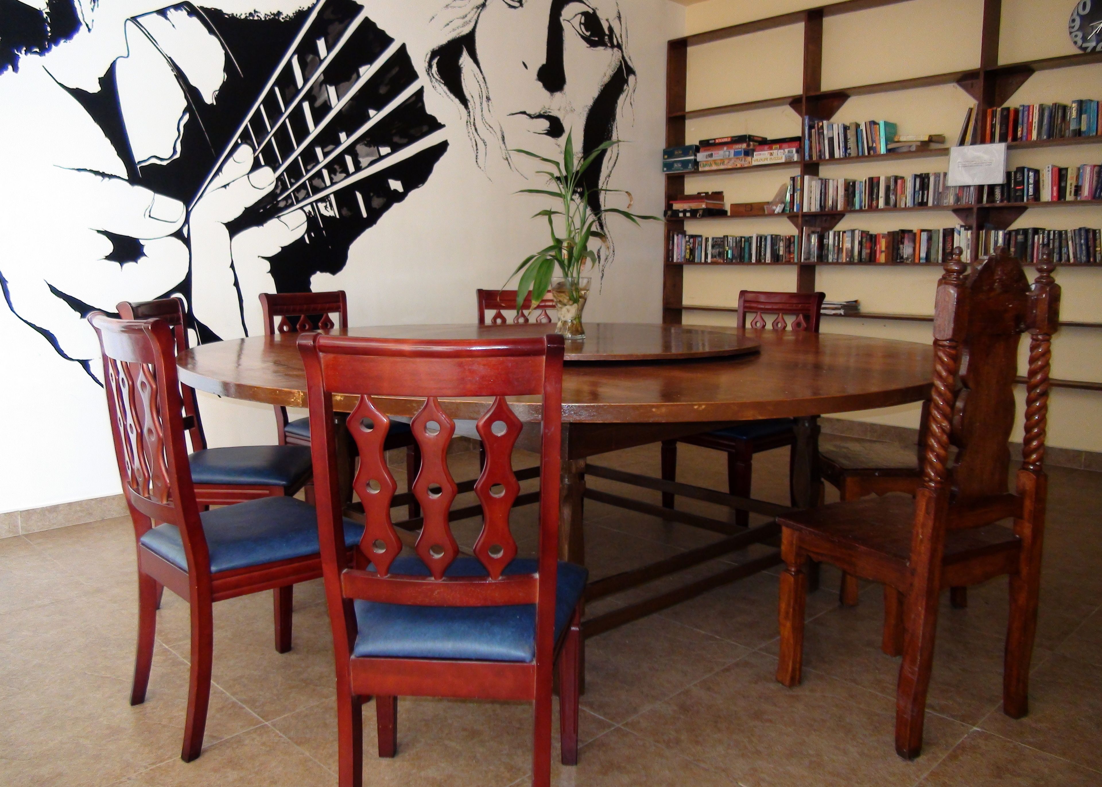 Un coin repas avec une table ronde en bois, quatre chaises dotées de coussins bleus, une bibliothèque remplie de livres et une grande fresque murale en noir et blanc.
