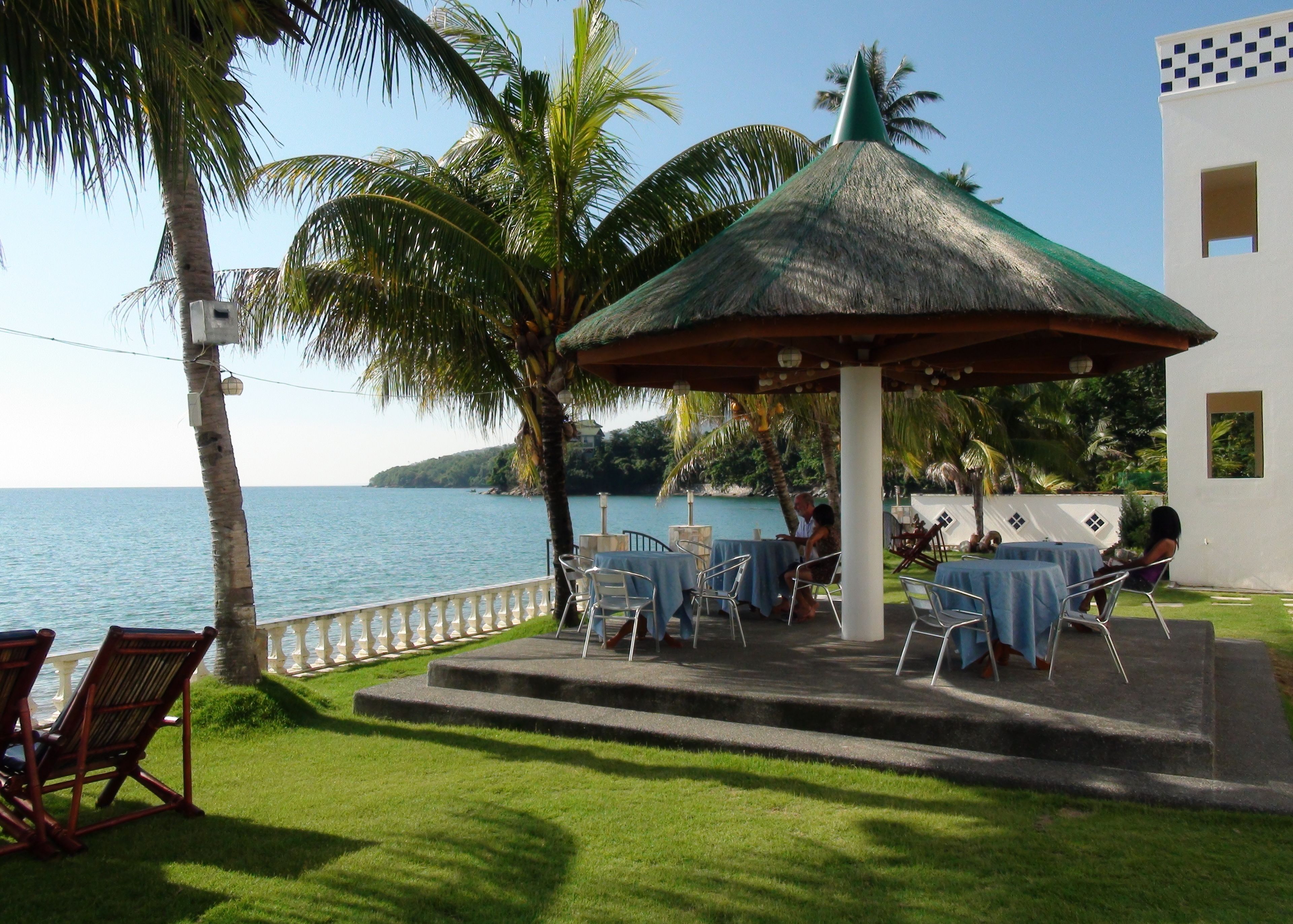 A covered outdoor seating area with tables and chairs, overlooking a body of water, surrounded by palm trees and a white building.
