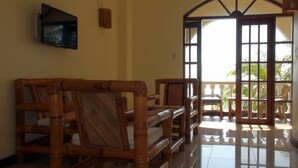 A room with bamboo furniture, a flat-screen TV, and a large window with a view of palm trees. - Squares Cafe and Apartments (Puerto Galera)