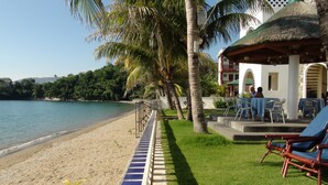 A beachside hotel with a thatched-roof structure, a pool area with lounge chairs, and a paved walkway along the beach. - Squares Cafe and Apartments (Puerto Galera)