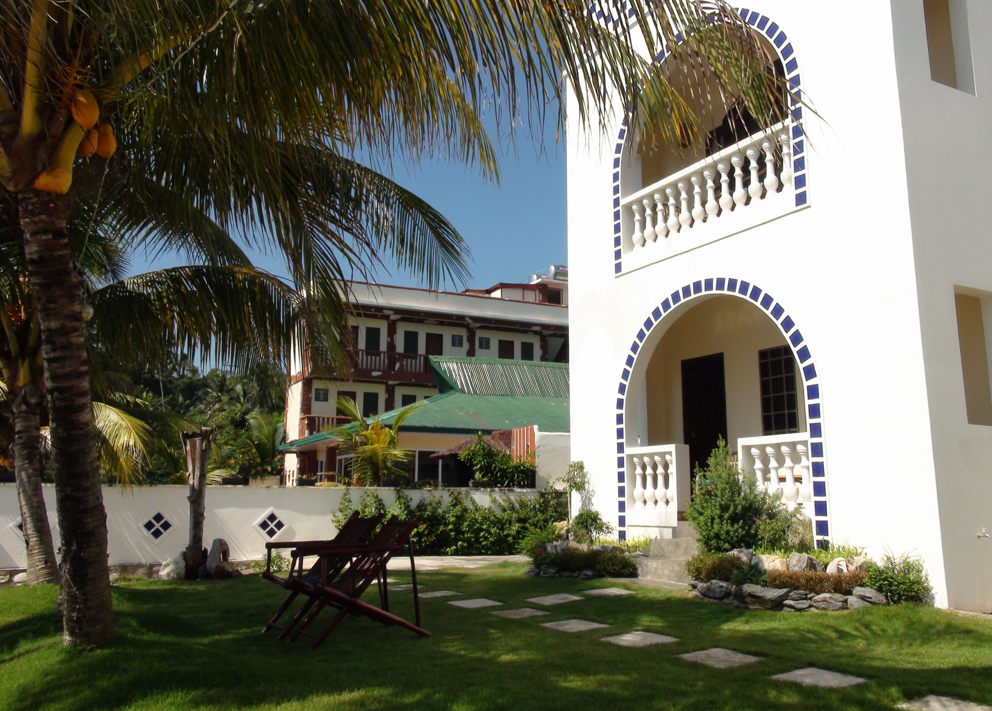 A two-story building with a balcony, a green roof, and a garden with palm trees and a swing set.