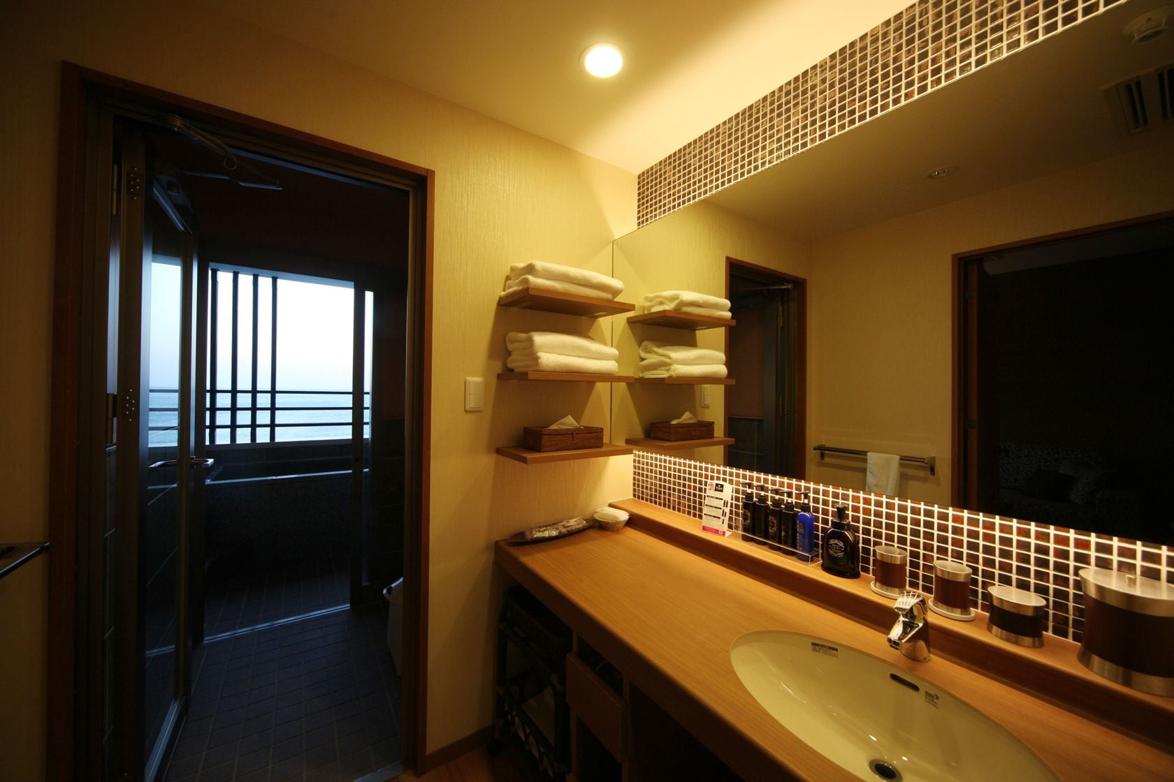 A hotel bathroom with a large mirror, wooden shelves with towels, a sink, and toiletries.