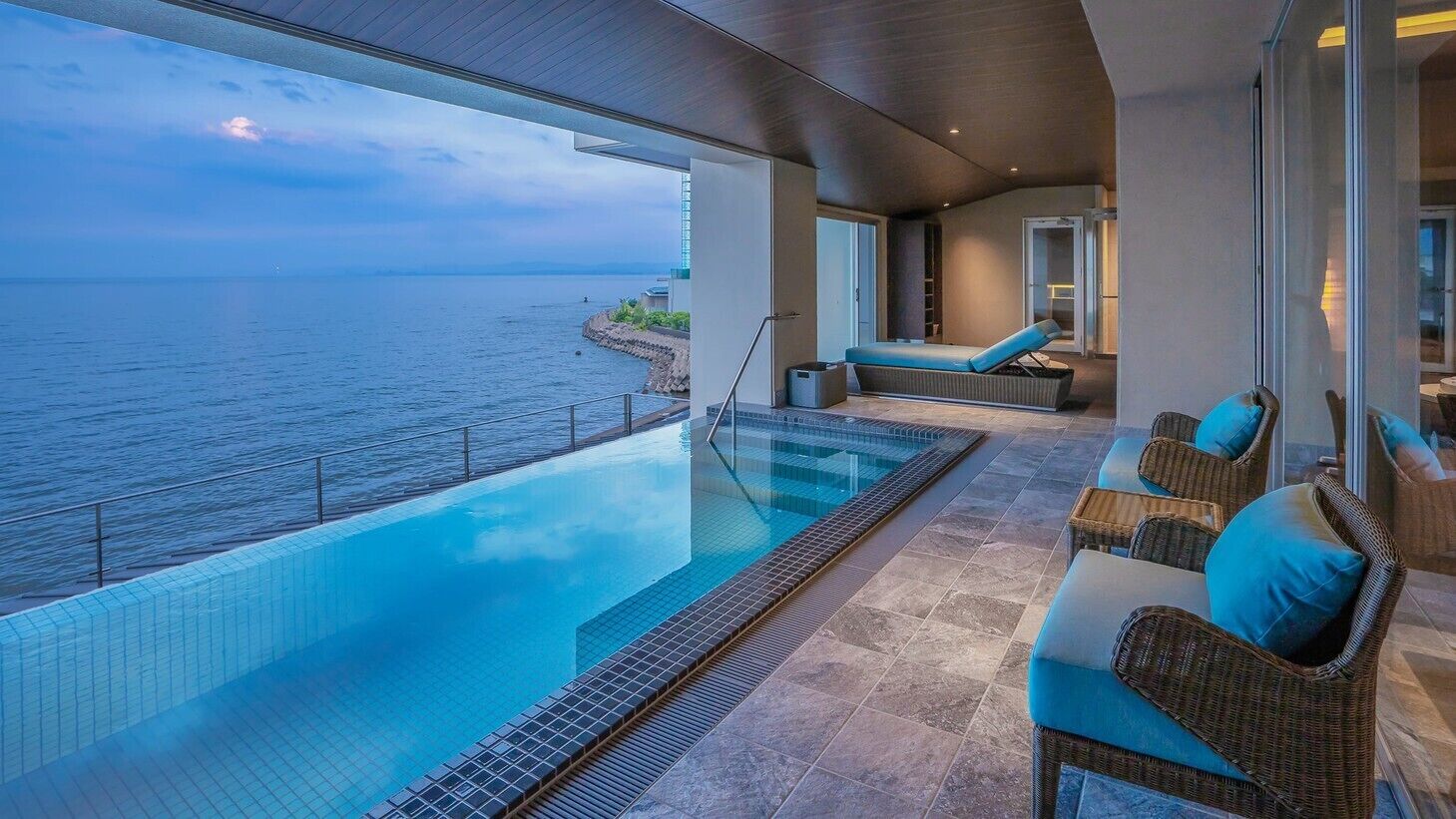 Een overloopzwembad met uitzicht op zee, een loungehoek met rieten meubels en een glazen deur.