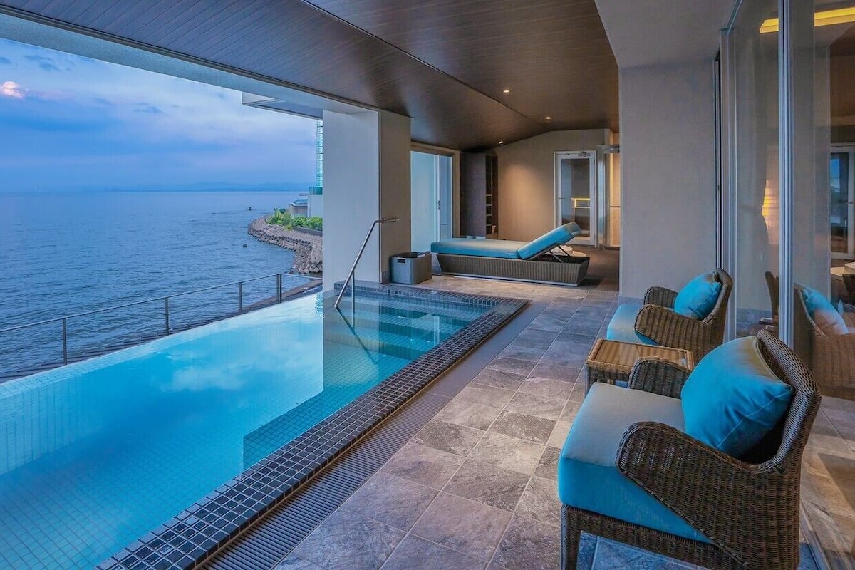 Une piscine à débordement offrant une vue sur la mer, un espace détente avec des meubles en osier et une porte à vitres.