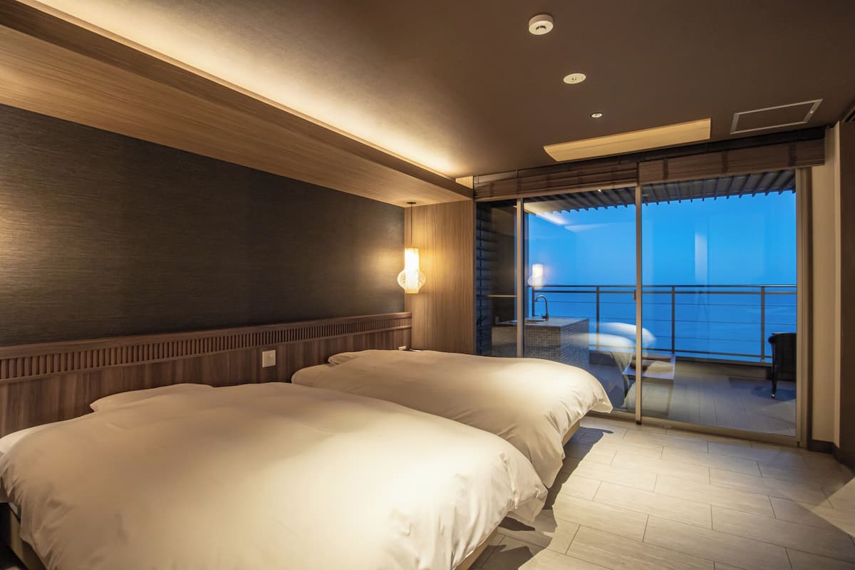 Quarto de hotel com duas camas, uma janela grande e varanda com vista para o mar.