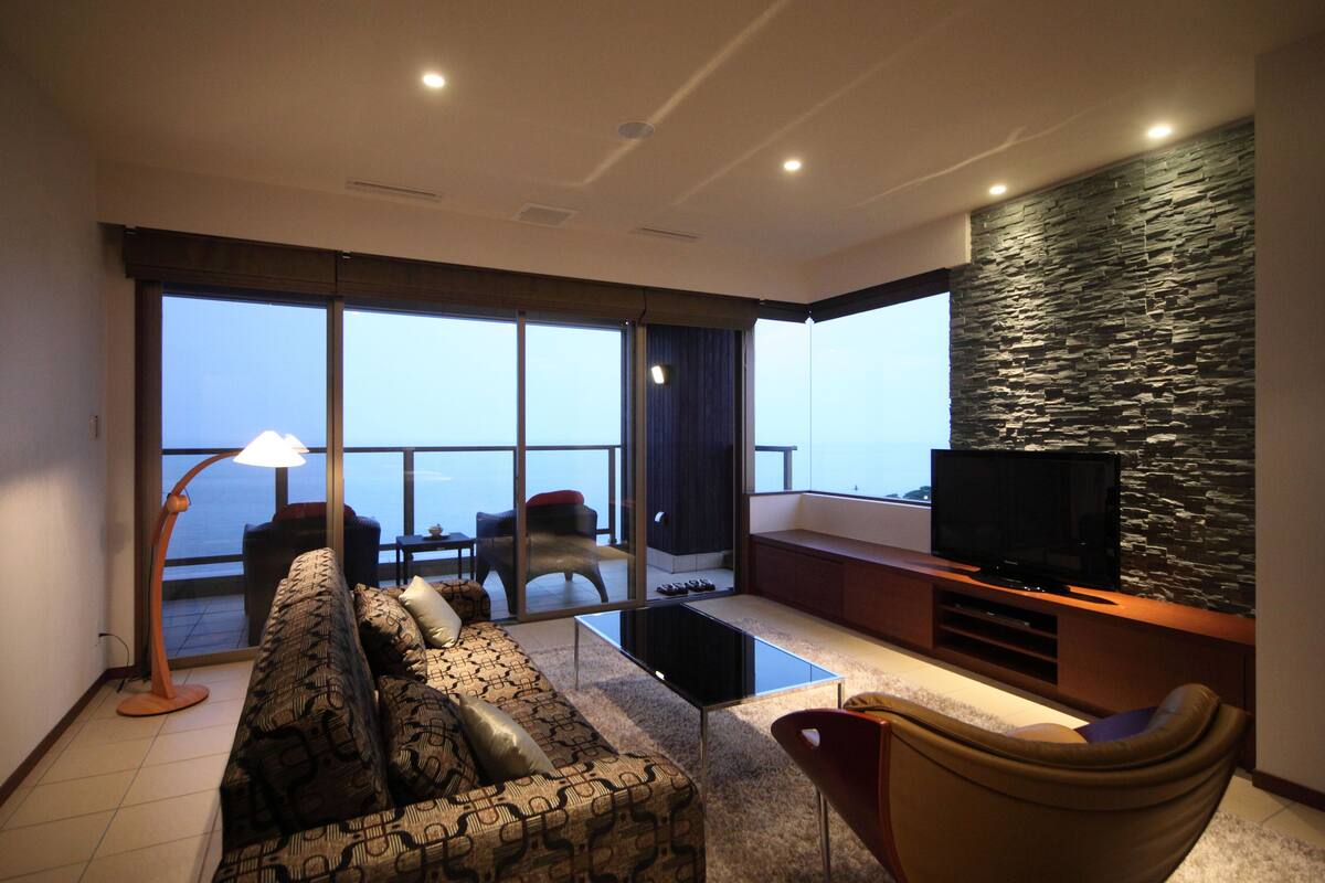 Ein modernes Wohnzimmer mit einer Couch, Sesseln, einem Couchtisch, einem Fernseher und einem Balkon mit Blick.