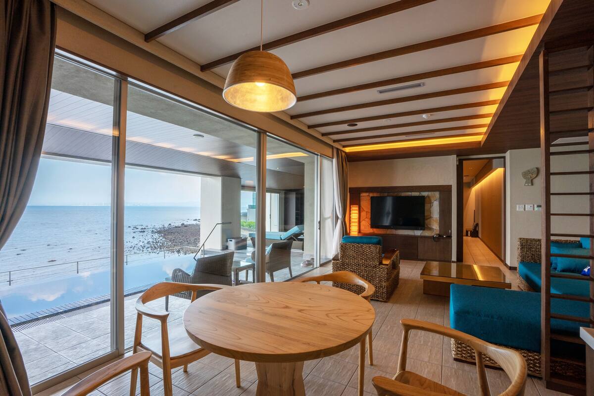 Ein modernes Interieur mit großem Fenster mit Meerblick, einem runden Holztisch und einer gemütlichen Sitzecke mit blauen Kissen.