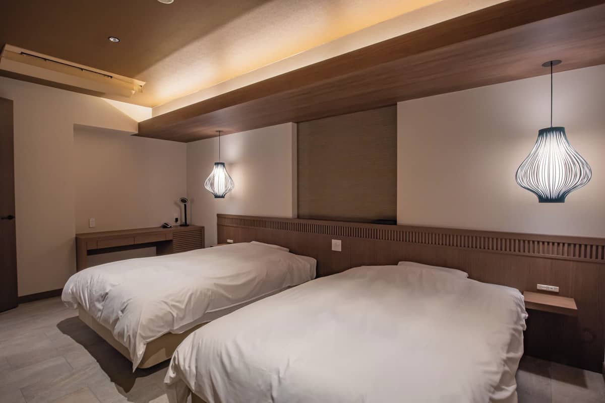 Quarto de hotel com duas camas, cabeceiras de madeira, uma mesa de cabeceira com abajur e um espelho grande na parede.