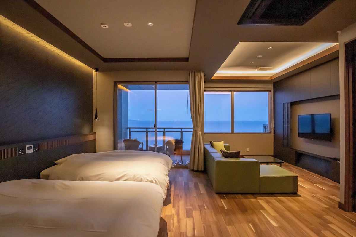 Quarto de hotel com varanda, cama, sofá, televisão e vista para o oceano.