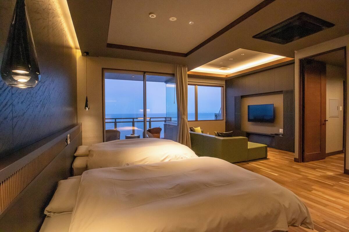 Quarto de hotel com duas camas, televisão, varanda com vista e área de estar.
