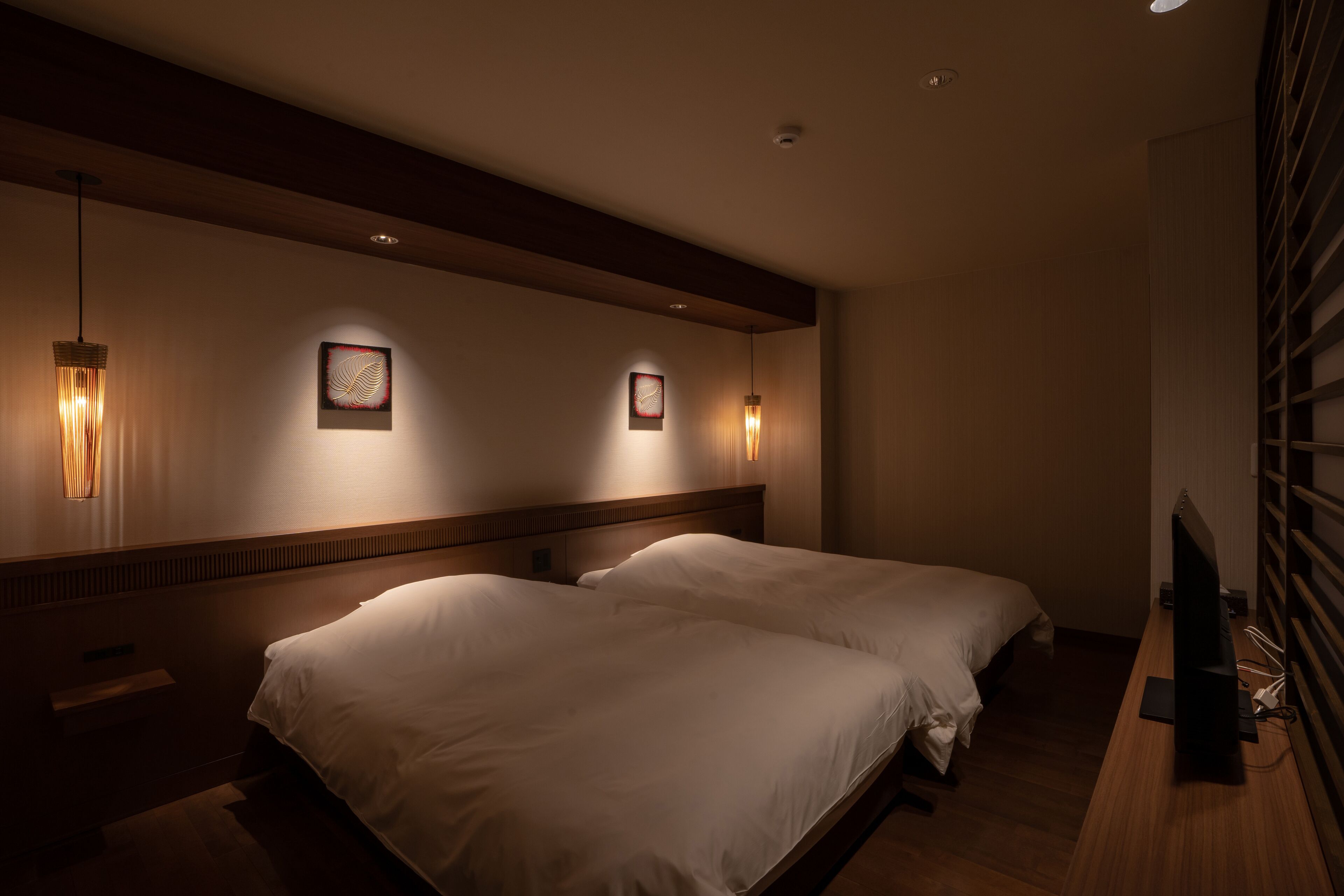 Une chambre d’hôtel avec deux lits, une tête de lit en bois, des œuvres d’art accrochées au mur et un téléviseur posé sur un bureau en bois.