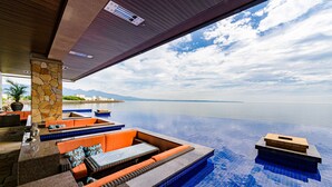 A modern infinity pool with a stone pillar and orange seating area, overlooking a calm sea and distant mountains.