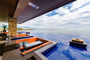 Une piscine à débordement moderne, agrémentée d’un pilier en pierre et d’un espace détente aux couleurs orangées, offrant une vue sur une mer calme et des montagnes lointaines.